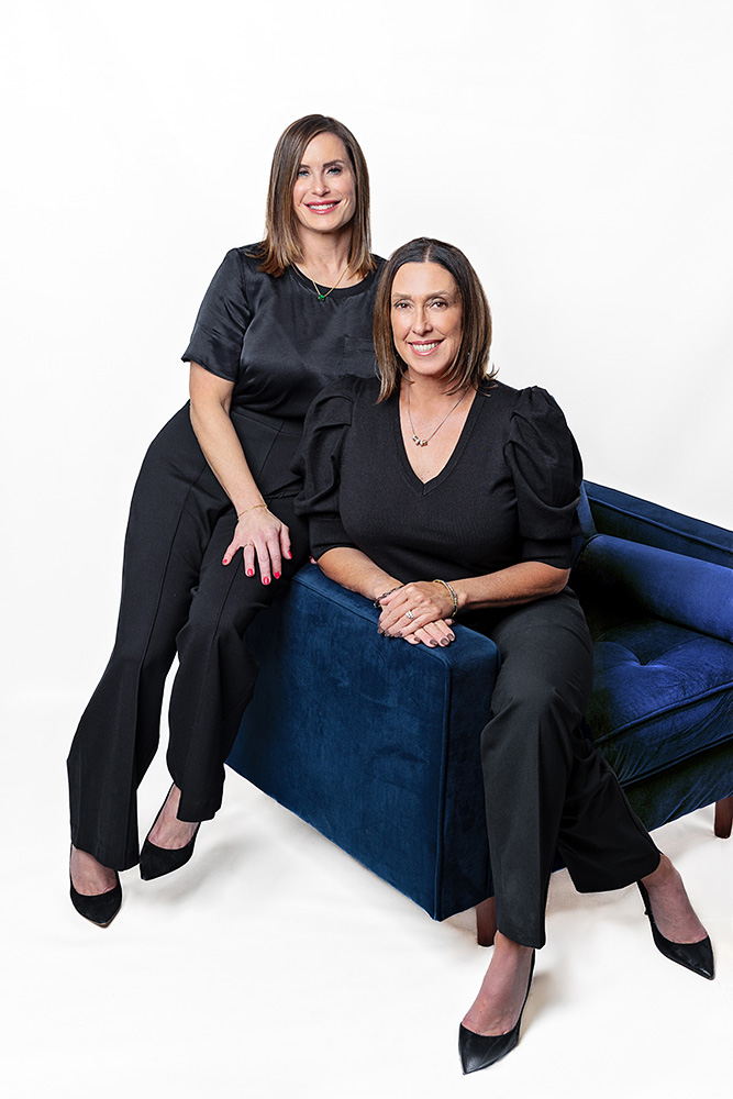 Two smiling female periodontists in black outfits pose on a blue couch at Mountainview Periodontics